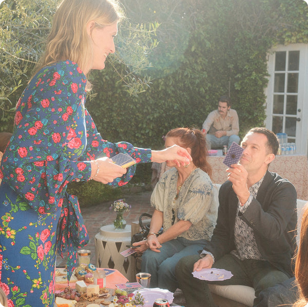 Smiling woman in a floral blue dress hands out tarot cards to friends during an outdoor gathering. People sit around a table with snacks, drinks, and flowers, enjoying the sunny atmosphere.