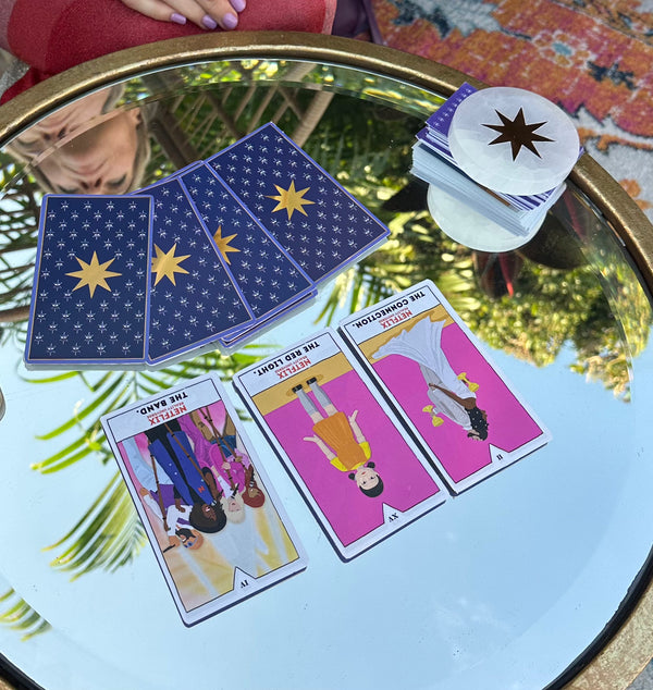 Tarot reading setup on a round glass table outdoors. Three tarot cards are laid face up showing illustrated figures with bright pink backgrounds, while several starry-backed cards remain face down. A stack of cards with a golden star design sits to the side. Reflections of palm leaves and a person’s face are visible in the glass surface.