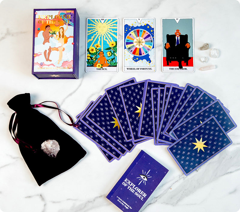 Explorer of the Soul tarot deck set displayed on a marble surface. The set includes a colorful tarot box, three revealed cards (The Sun, Wheel of Fortune, and The Emperor), a spread of starry-backed cards, a purple guidebook, a black pouch with a crystal, and several small clear and rose quartz stones.