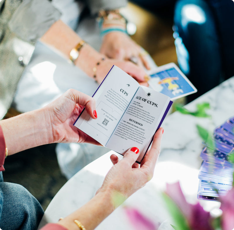 Close-up of a person holding a tarot guidebook open to the 'Ace of Cups' page, with another person in the background holding a tarot card. The scene is set at a marble table with a stack of tarot cards and flowers nearby.