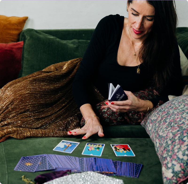 Woman sitting on a green sofa with colorful pillows, holding a tarot guidebook while reading cards laid out on the cushion in front of her. Three tarot cards are face up, with the rest of the deck spread nearby. She wears a black top, gold skirt, and jewelry, creating a cozy, reflective atmosphere.