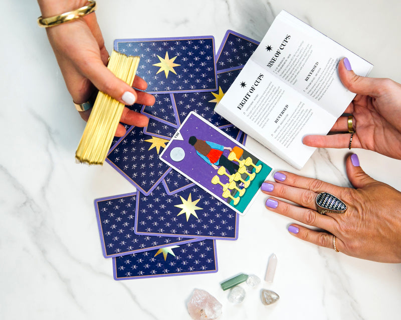 Person holding tarot cards and a deck with crystals and a book on a white surface
