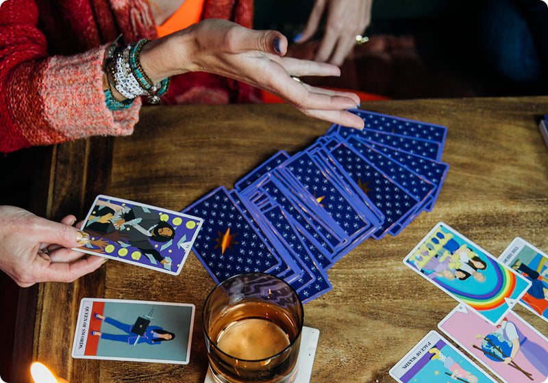 Two people engaged in a tarot reading at a wooden table. One hand holds a tarot card while others are spread face down with starry blue backs. Several cards are revealed, including colorful illustrations, next to a glass of amber-colored drink and a lit candle, creating a warm, cozy atmosphere.