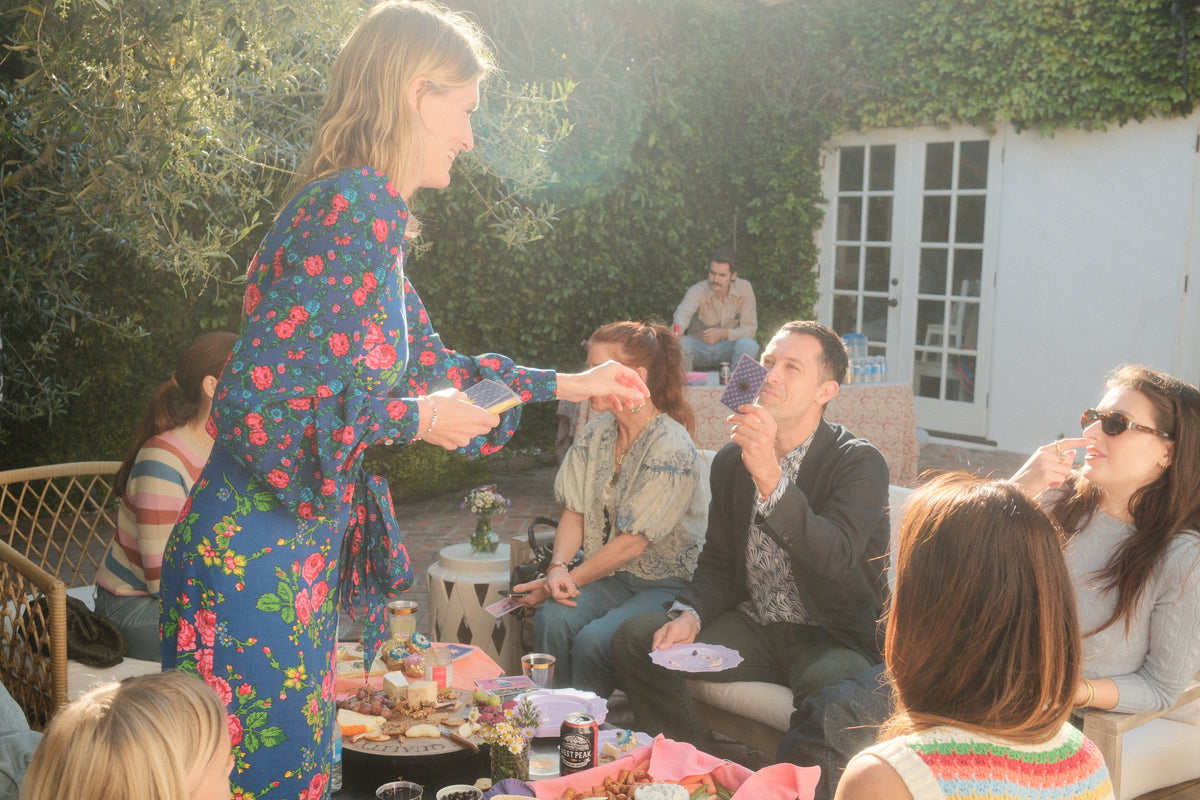 Woman in floral dress addressing a group of people outdoors in a garden setting.